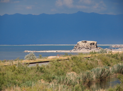 [A rock outcroppping near the water's edge appears to be covered in salt. There is sage growing on the land. In the background covering more than a third of the image is the dark blue shadow outline of the Wasatch Mountains. There are some puffy clouds in the sky above the distant mountains.]